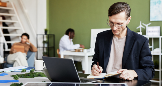 Young businessman making notes in notebook while working at table with laptop and solar panel