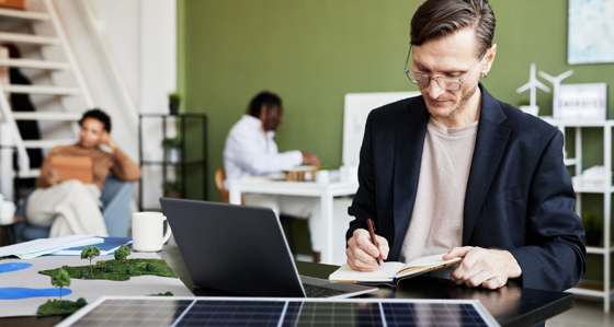Young businessman making notes in notebook while working at table with laptop and solar panel