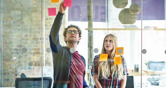 A man writing data on a perspex wall with colleague watching